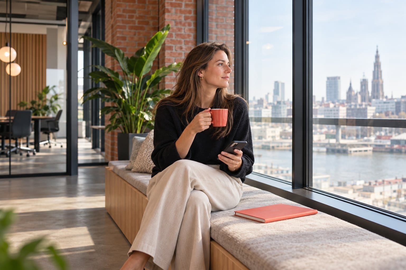 Een recruiter met een koffie en haar gsm kijkt vanop een zitbank aan het raam uit over Antwerpen
